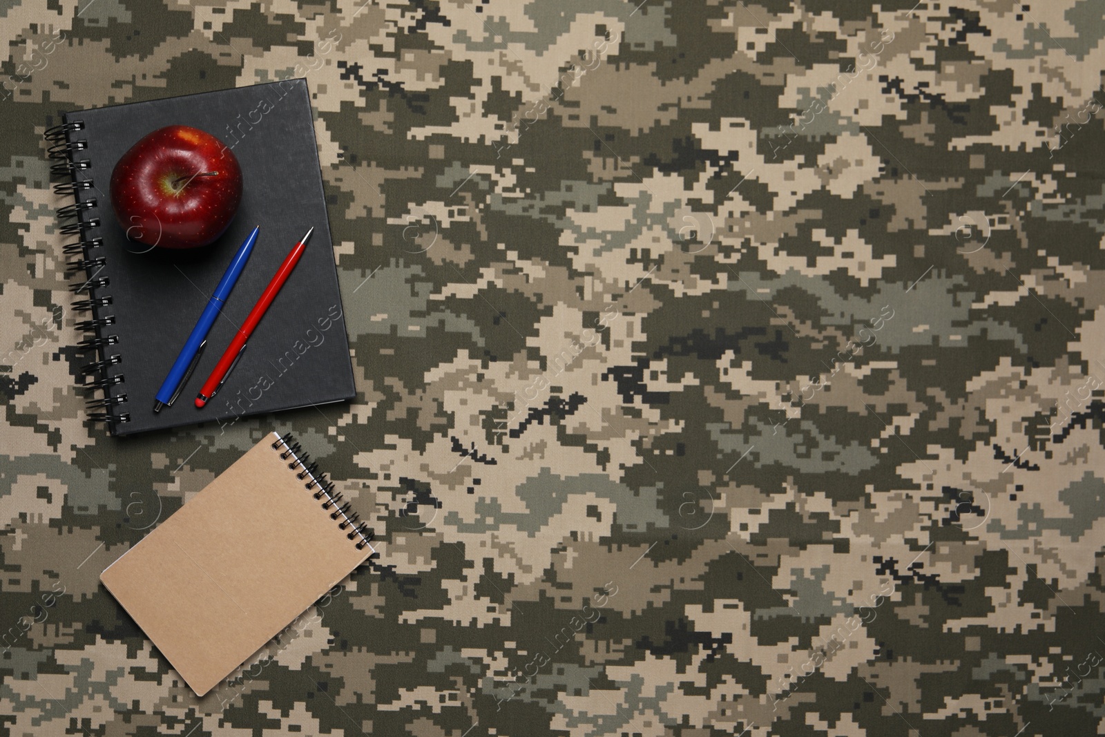 Photo of Notebooks, apple and pens on camouflage fabric, flat lay with space for text. Military education