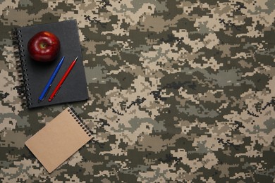 Photo of Notebooks, apple and pens on camouflage fabric, flat lay with space for text. Military education