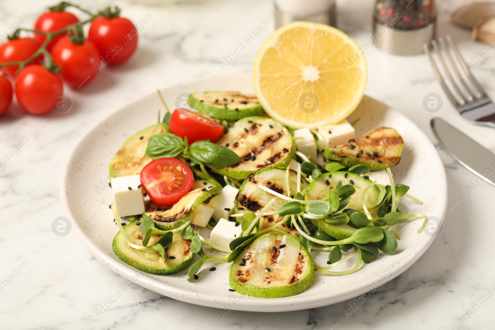 Photo of Delicious grilled zucchini slices served with feta cheese, tomatoes and lemon on white marble table, closeup