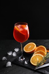 Photo of Glass of tasty Aperol spritz cocktail with orange slices, ice cubes and rosemary on table against black background