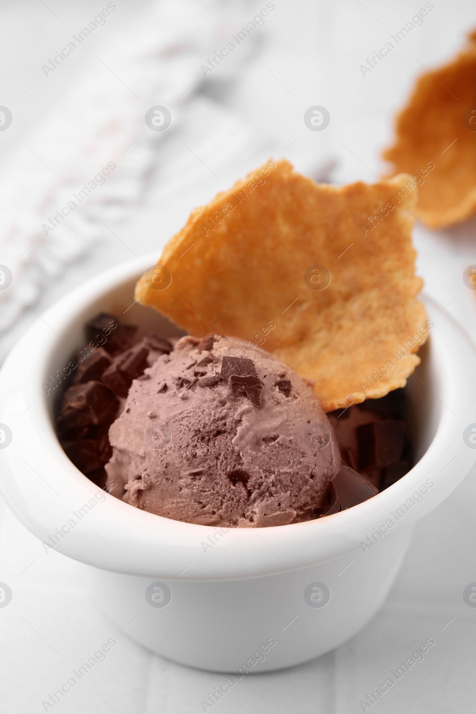 Photo of Tasty chocolate ice cream and piece of waffle cone in bowl on white table, closeup