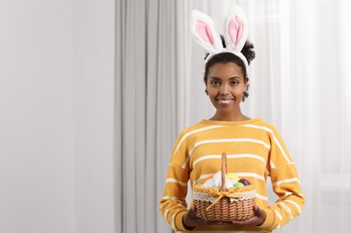 Photo of Happy African American woman in bunny ears headband holding wicker basket with Easter eggs indoors, space for text