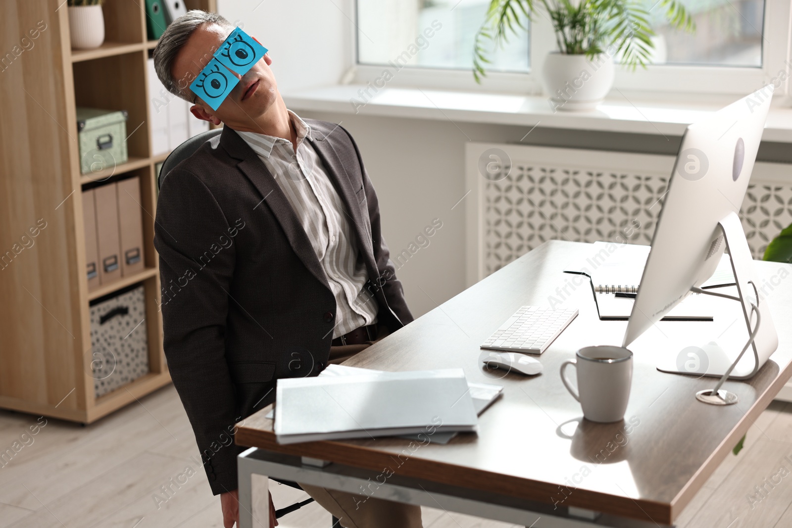 Photo of Man with fake eyes painted on sticky notes snoozing at workplace in office