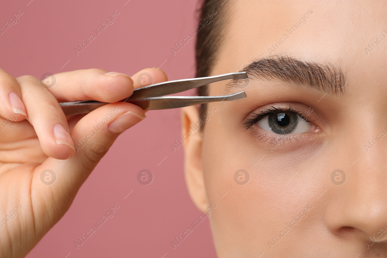 Photo of Eyebrow correction. Young woman with tweezers on pink background, closeup