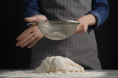 Photo of Making bread. Woman sifting flour over dough at table on dark background, closeup