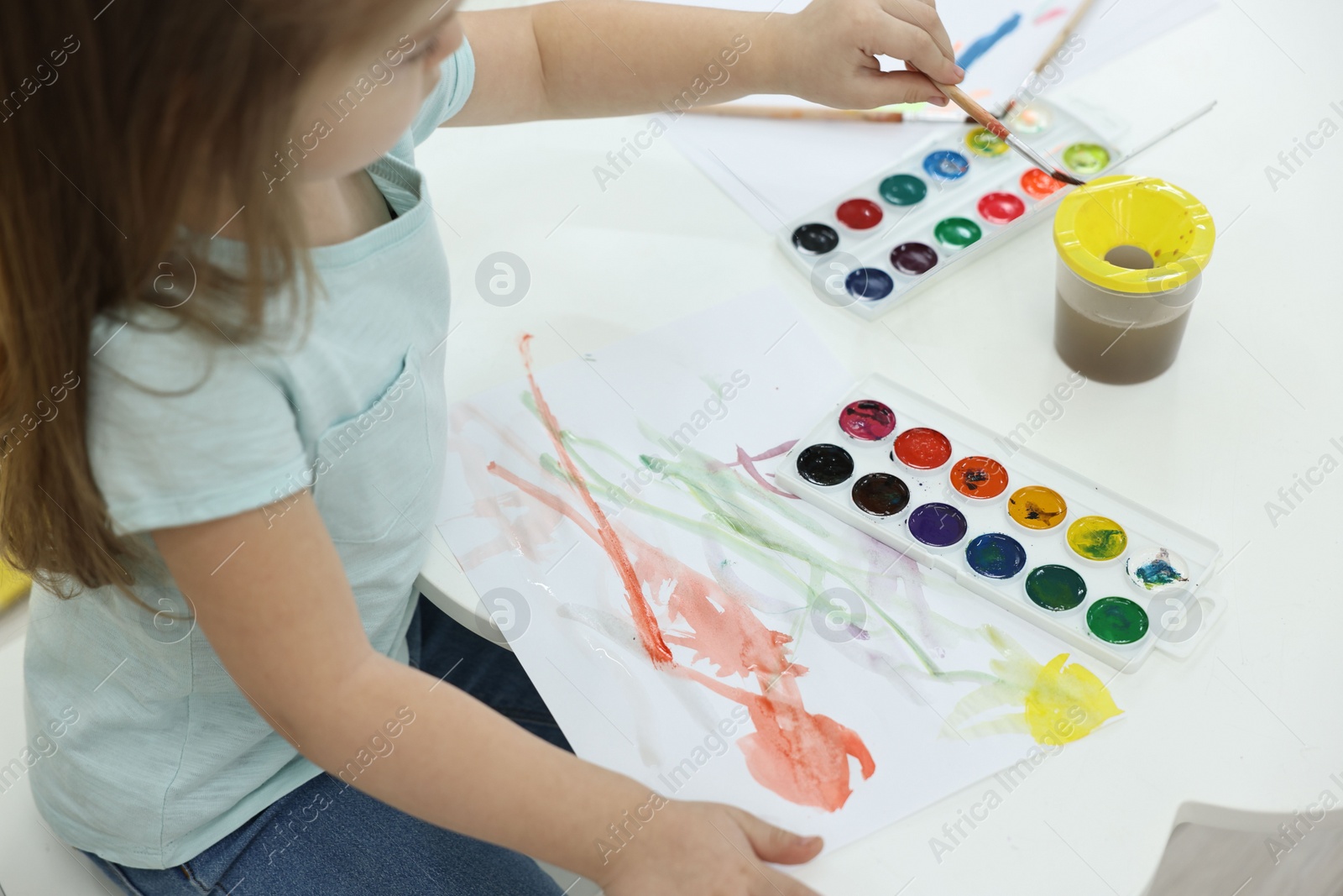 Photo of Little girl painting with brush and watercolor at white table, closeup