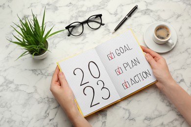 Photo of Woman holding notebook with text 2023 Goal, Plan, Action at white marble table, top view. New Year resolutions