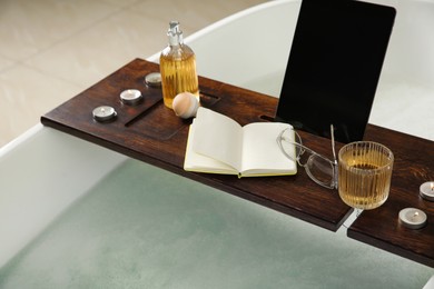 Photo of Wooden bath tray with candles, tablet, book and glass of drink on tub, closeup