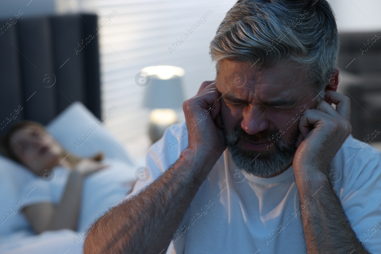 Photo of Irritated man covering his ears in bed at home, space for text. Problem with snoring wife