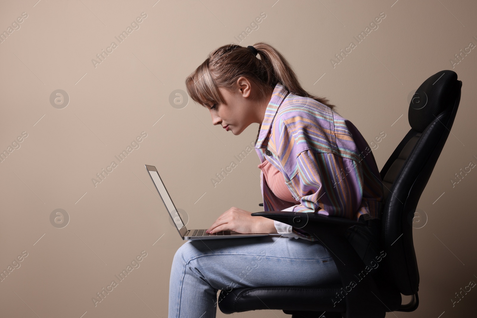 Photo of Young woman with poor posture using laptop while sitting on chair against beige background