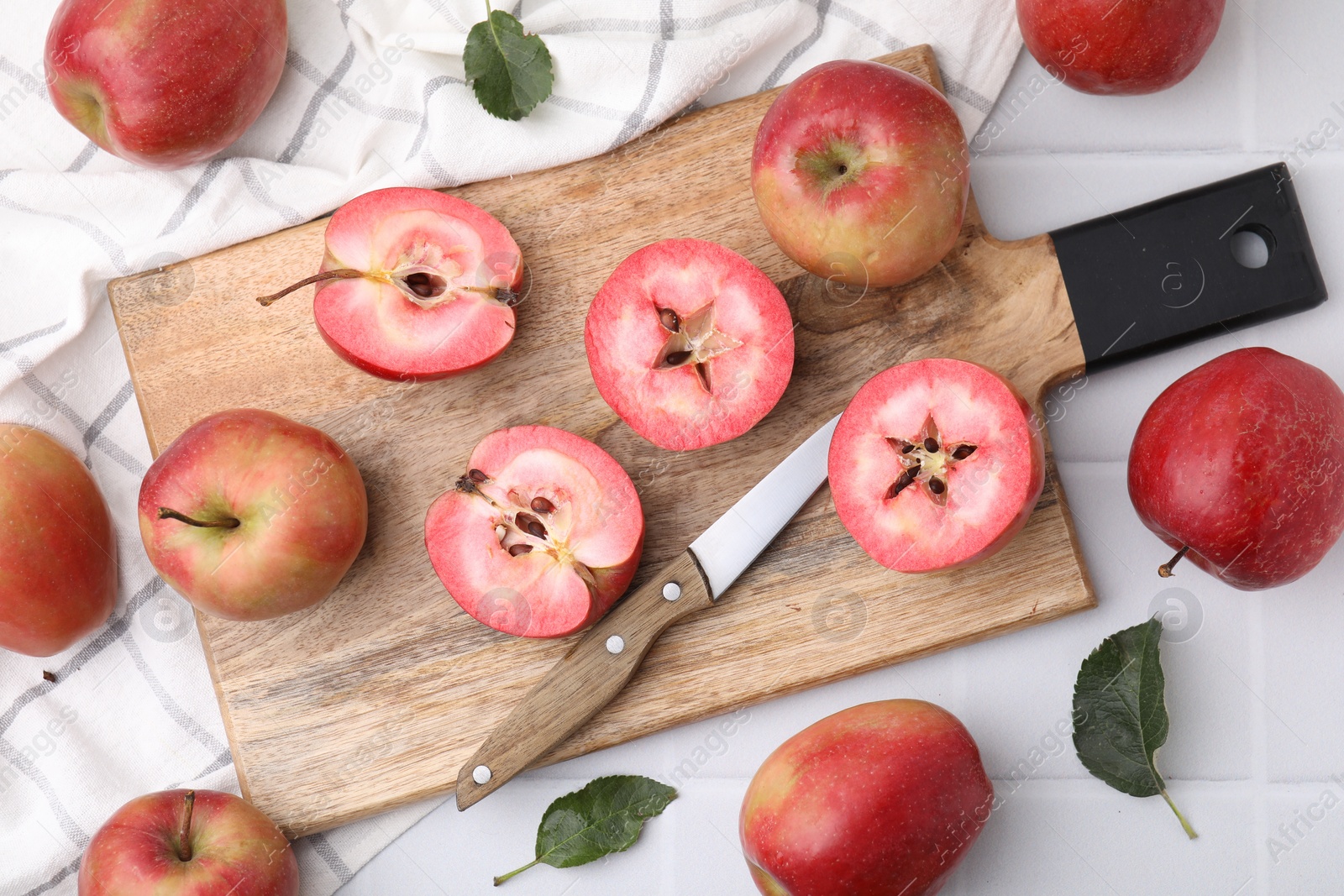 Photo of Tasty apples with red pulp, leaves and knife on white tiled table, flat lay