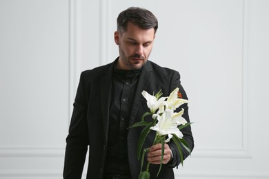 Sad man with lily flowers near white wall. Funeral ceremony