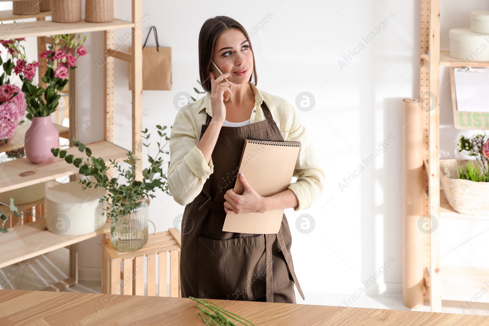 Photo of Florist with notebook talking on smartphone in workshop