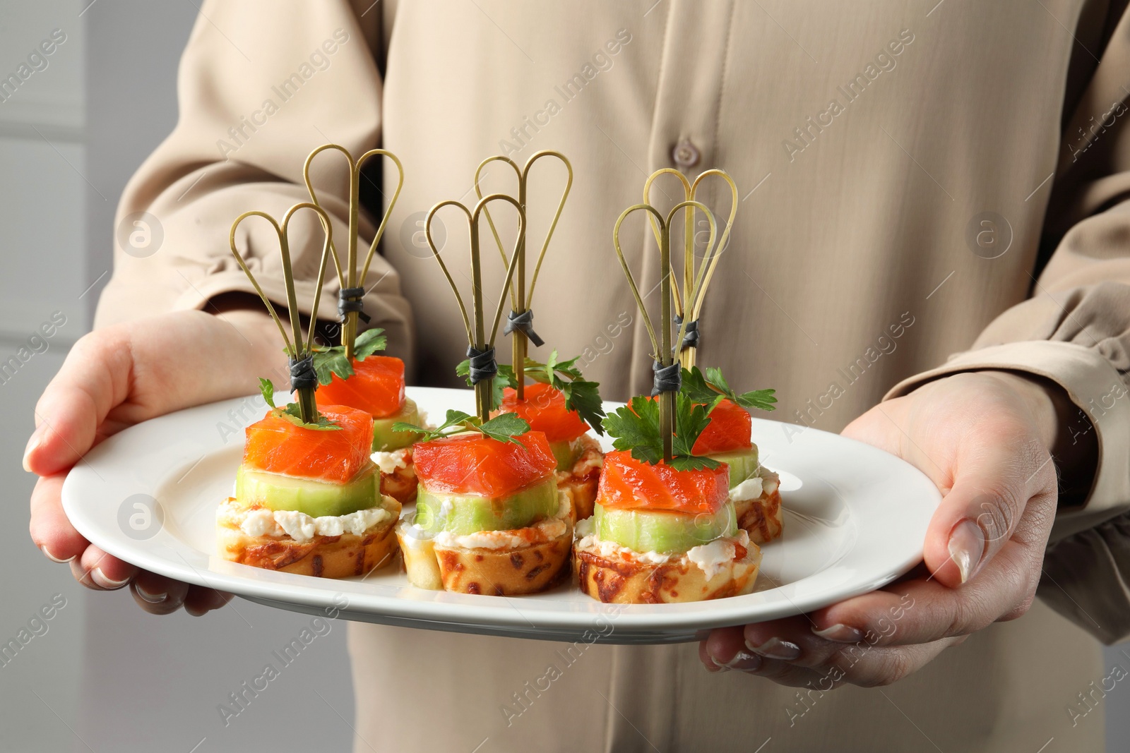 Photo of Woman holding tasty canapes with salmon, cucumber, bread and cream cheese on grey background, closeup