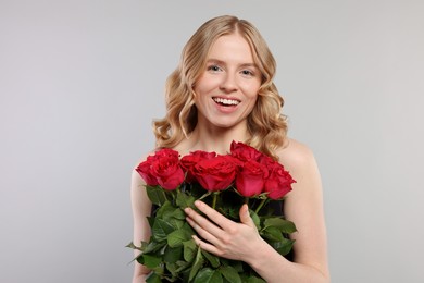 Beautiful woman with blonde hair holding bouquet of red roses on light grey background