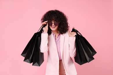 Happy young woman with shopping bags and stylish sunglasses on pink background