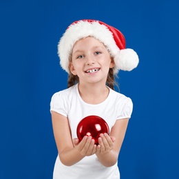 Happy little child in Santa hat holding Christmas ball on blue background