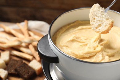 Dipping piece of bread into fondue pot with melted cheese on blurred background, closeup