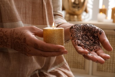 Woman with henna tattoo holding burning candle indoors, closeup. Traditional mehndi ornament