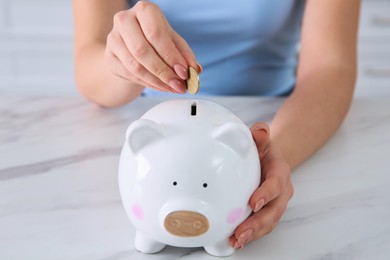 Photo of Young woman putting coin into piggy bank at table indoors, closeup. Money savings