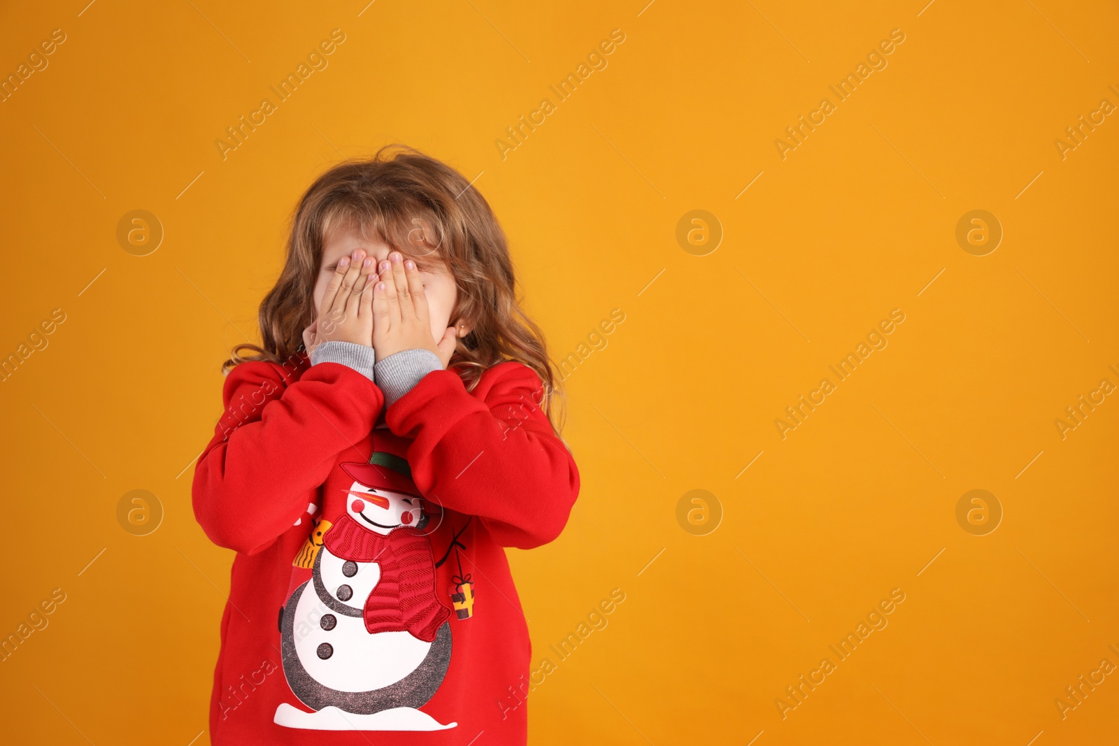 Photo of Cute little girl in red Christmas sweater covering her face against orange background. Space for text