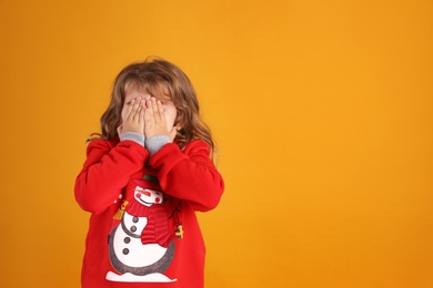 Cute little girl in red Christmas sweater covering her face against orange background. Space for text