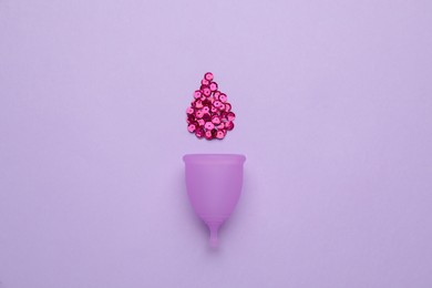 Menstrual cup near drop made of pink sequins on violet background, flat lay