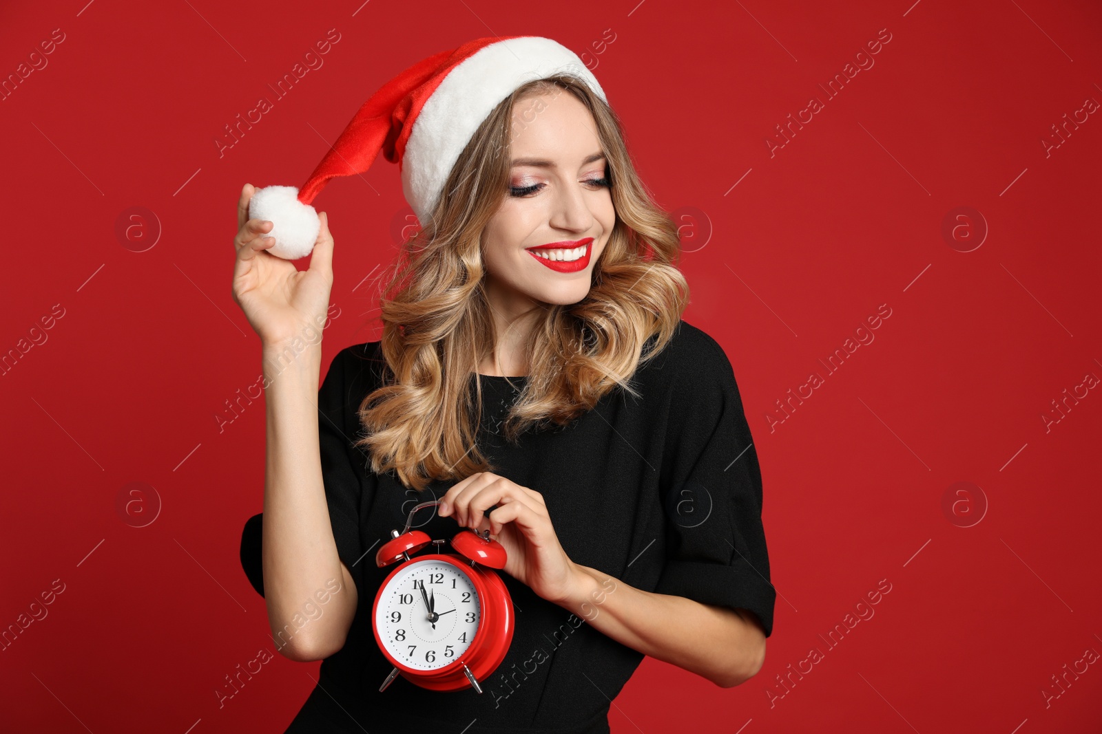 Photo of Happy young woman wearing Santa hat with alarm clock on red background. Christmas time