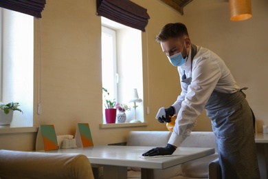 Photo of Waiter cleaning table with rag and detergent in restaurant. Surface treatment during coronavirus quarantine