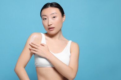 Beautiful young Asian woman applying body cream onto shoulder on light blue background, space for text