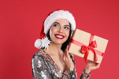 Woman in silver dress and Santa hat holding Christmas gift on red background