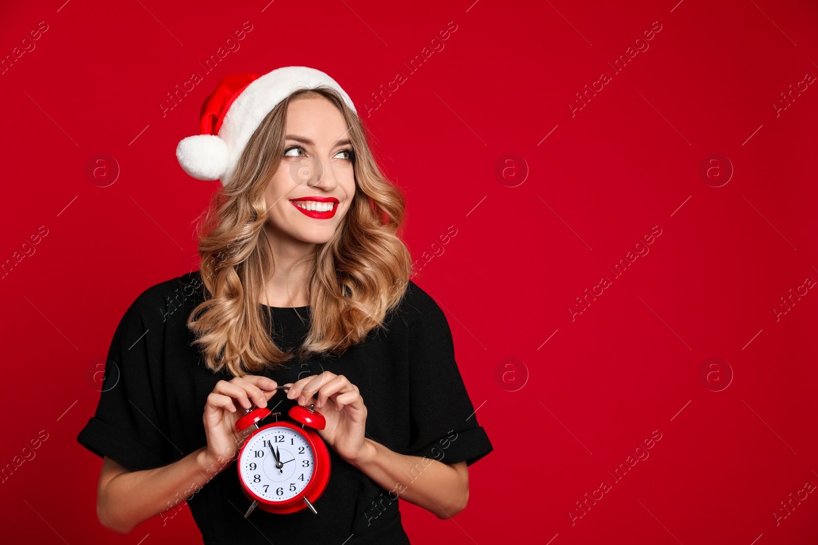 Photo of Happy young woman wearing Santa hat with alarm clock on red background, space for text. Christmas time