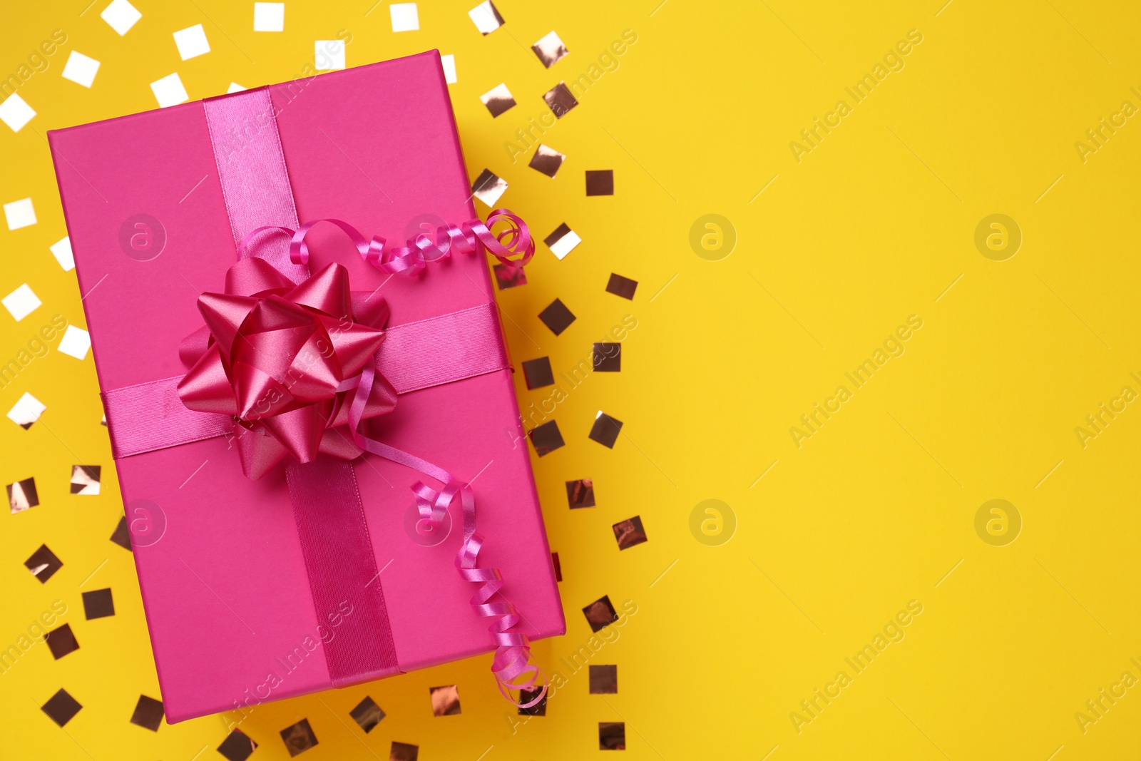 Photo of Pink gift box and shiny confetti on yellow background, top view. Space for text