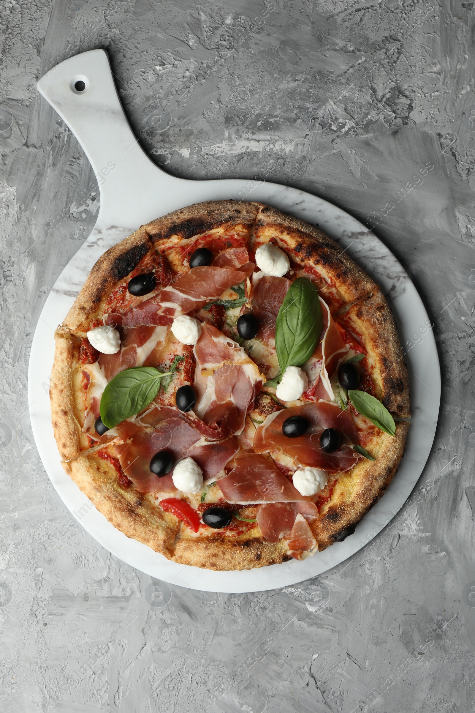 Photo of Tasty pizza with cured ham, olives, sun-dried tomato and basil on gray textured table, top view