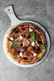 Photo of Tasty pizza with cured ham, olives, sun-dried tomato and basil on gray textured table, top view