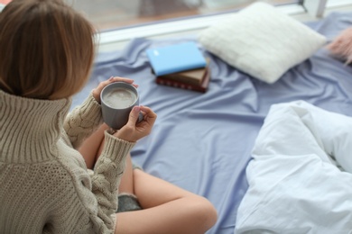 Photo of Beautiful young woman in knitted sweater sitting with cup of coffee at home. Winter atmosphere