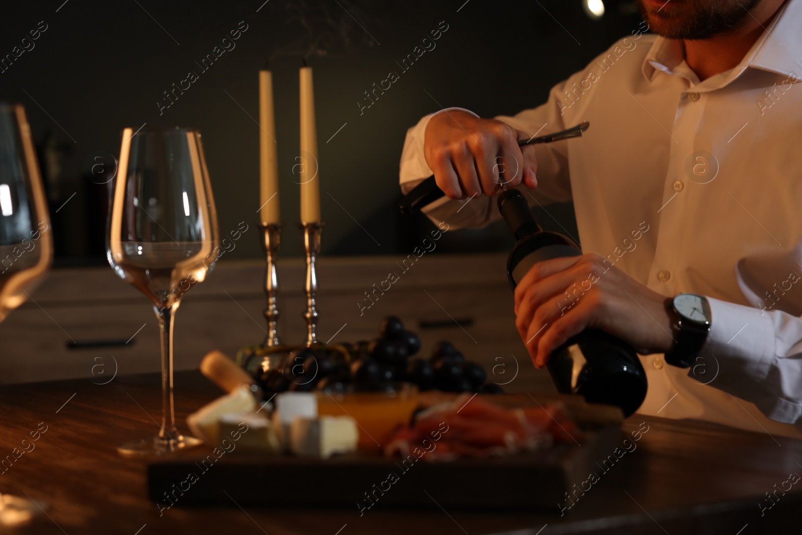 Photo of Romantic dinner. Man opening wine bottle with corkscrew at table indoors, closeup