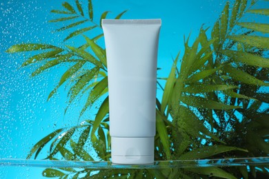 Tube with moisturizing cream and palm leaves on light blue background, view through wet glass