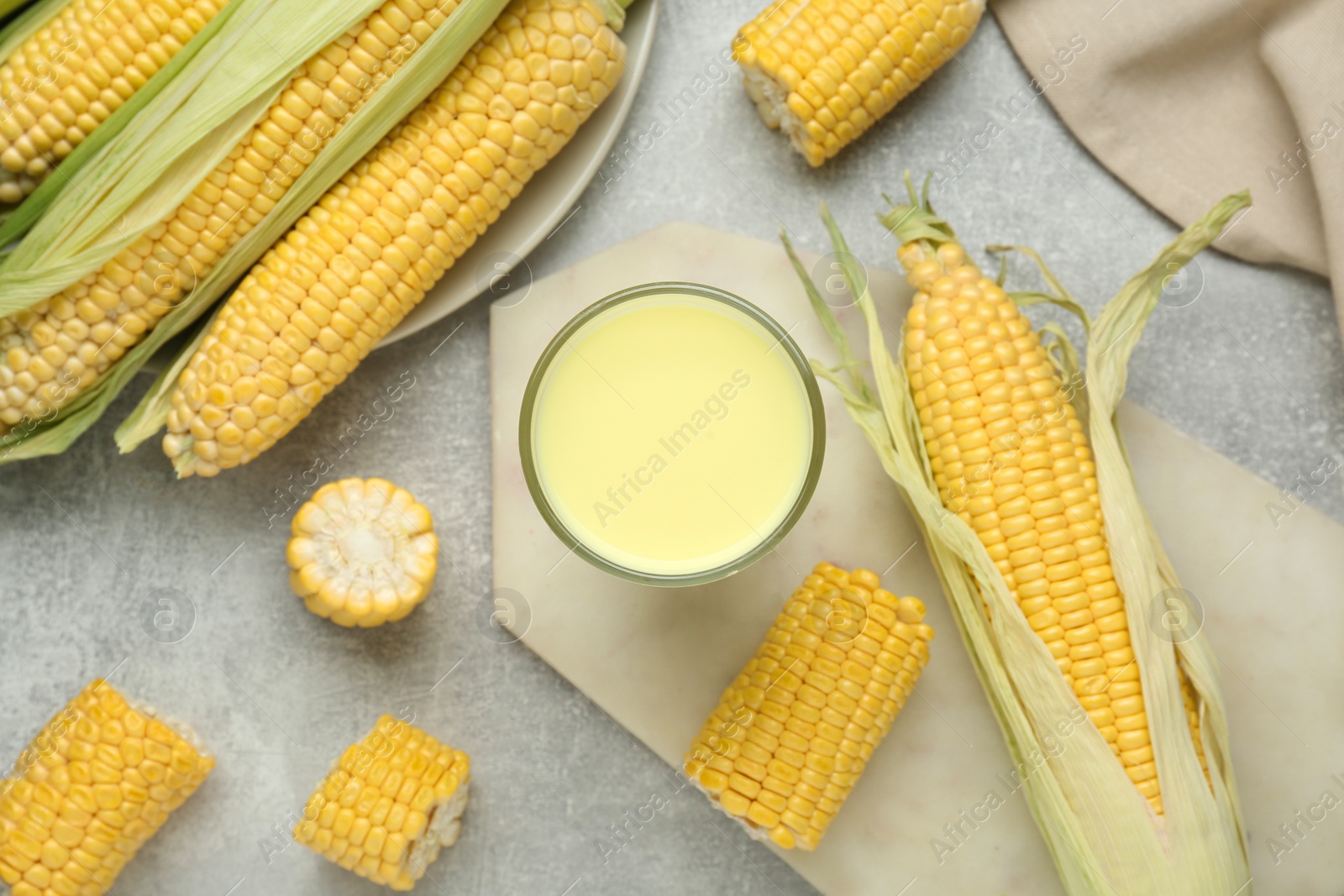 Photo of Tasty fresh corn milk in glass and cobs on light grey table, flat lay