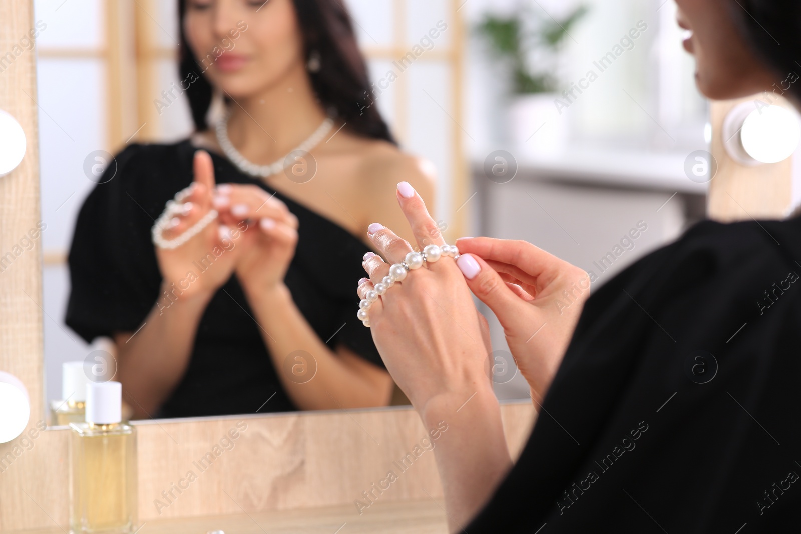 Photo of Young woman trying on elegant pearl bracelet near mirror indoors, closeup