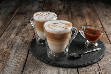 Glass cups of caramel macchiato and gravy boat with syrup served on wooden table