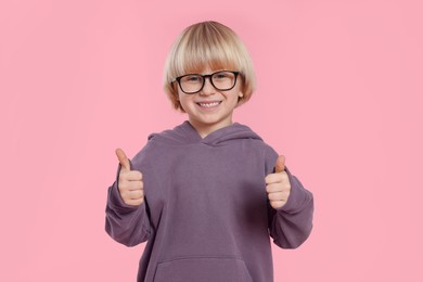 Cute little boy in glasses showing thumbs up on pink background