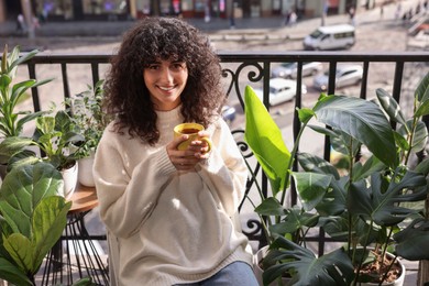 Photo of Beautiful young woman with cup of tea surrounded by houseplants on balcony