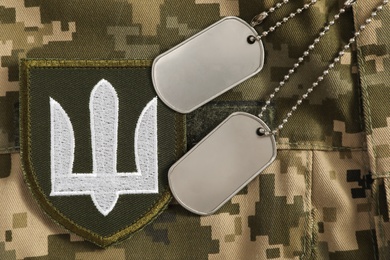 Photo of MYKOLAIV, UKRAINE - SEPTEMBER 26, 2020: Metal military ID tags and Ukrainian army patch on camouflage background, flat lay