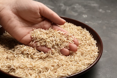 Photo of Woman holding grains near plate with brown rice on table, closeup