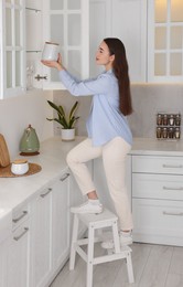 Photo of Woman on ladder putting white jar into cupboard at home