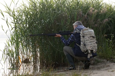 Man aiming with hunting rifle near lake outdoors. Space for text