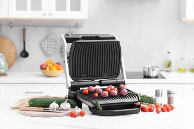 Photo of Electric grill with meatballs, tomatoes and vegetables on white marble table in kitchen