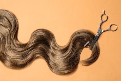 Photo of Lock of brown wavy hair and thinning scissors on color background, flat lay