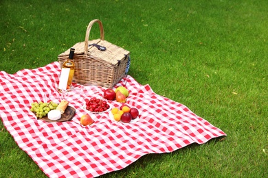 Photo of Picnic basket with products and bottle of wine on checkered blanket in garden